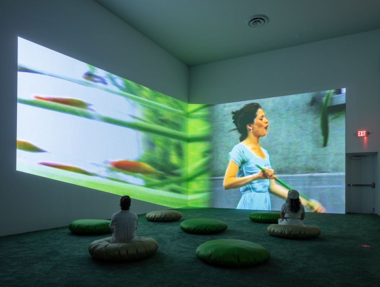 Installation view of Pipilotti Rist: Big Heartedness, Be My Neighbor at The Geffen Contemporary at MOCA, where two people are individually sitting on padded cushions on the floor watching a video projected on the wall showing a dark-haired woman dressed in blue is holding a green broom